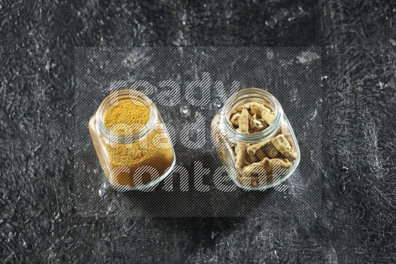 2 glass spice jars full of turmeric powder and dried whole fingers on textured black flooring