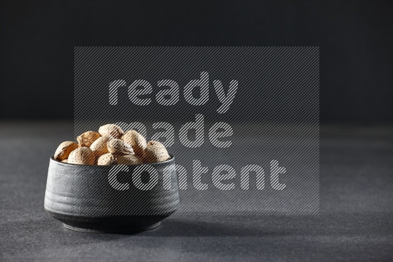A black pottery bowl full of almonds on a black background in different angles
