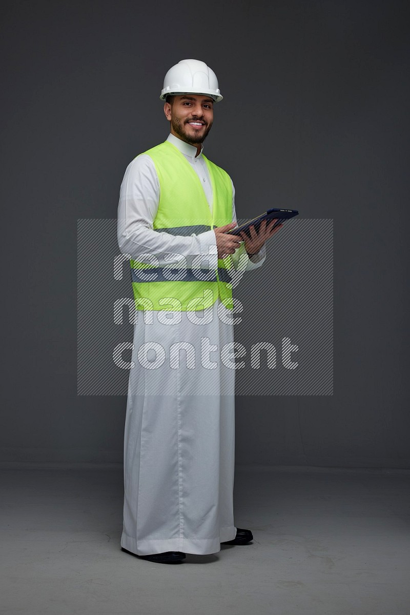 A Saudi man wearing Thobe with a yellow safety vest and white helmet standing and using his tablet different angles eye level on a gray background