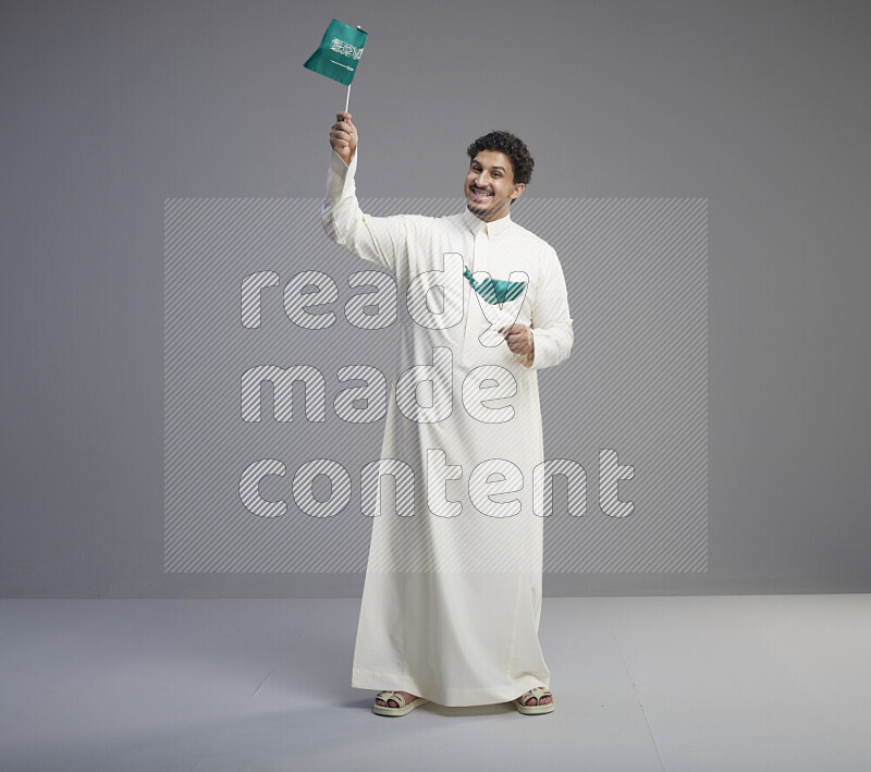 A Saudi man standing wearing thob holding small Saudi flag on gray background