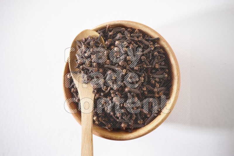 A wooden bowl and a wooden spoon full of cloves on a white flooring