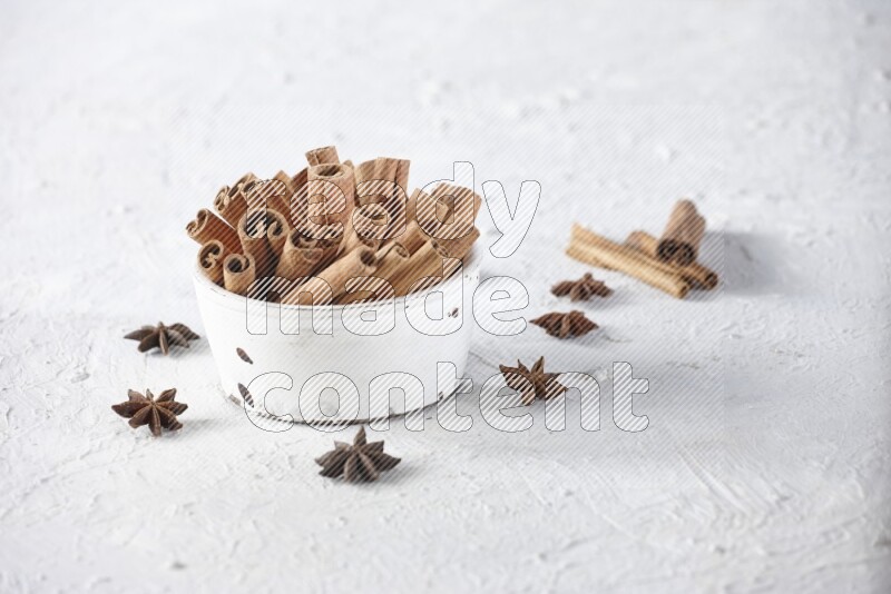 White bowl full of cinnamon sticks surrounded by star anis on a textured white background in different angles