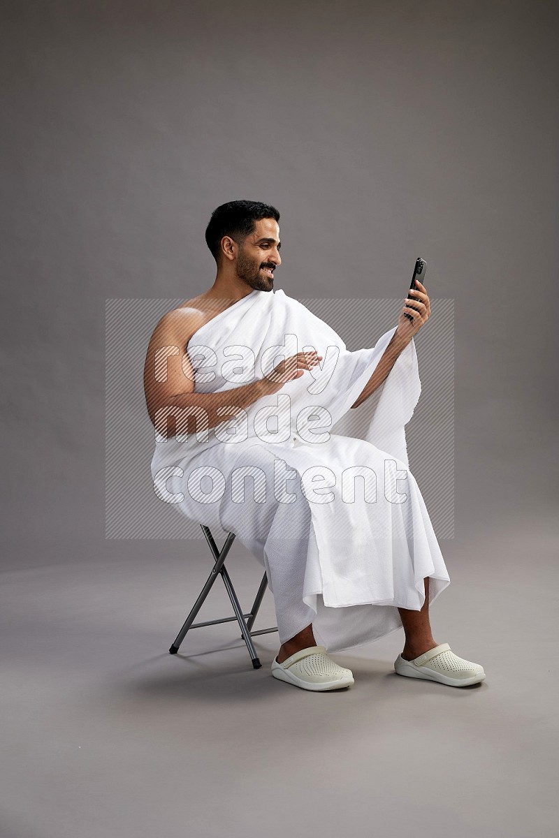 A man wearing Ehram sitting on chair taking selfie on gray background