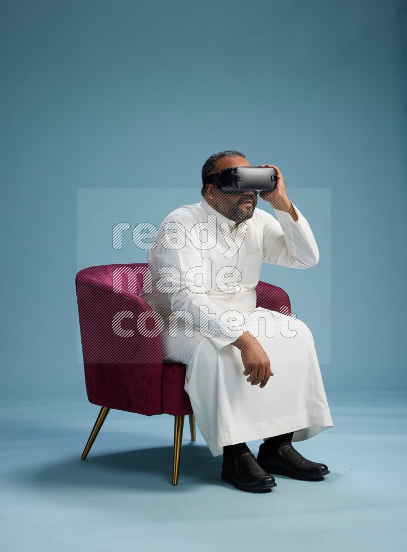 Saudi Man without shimag sitting on chair  gaming with VR on blue background