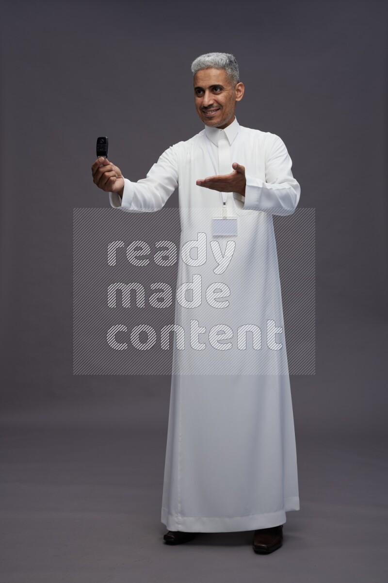 Saudi man wearing thob with neck strap employee badge standing holding key car on gray background