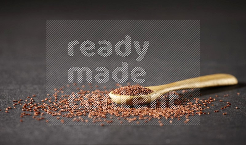 A wooden spoon full of garden cress seeds and seeds spread beside it on a black flooring