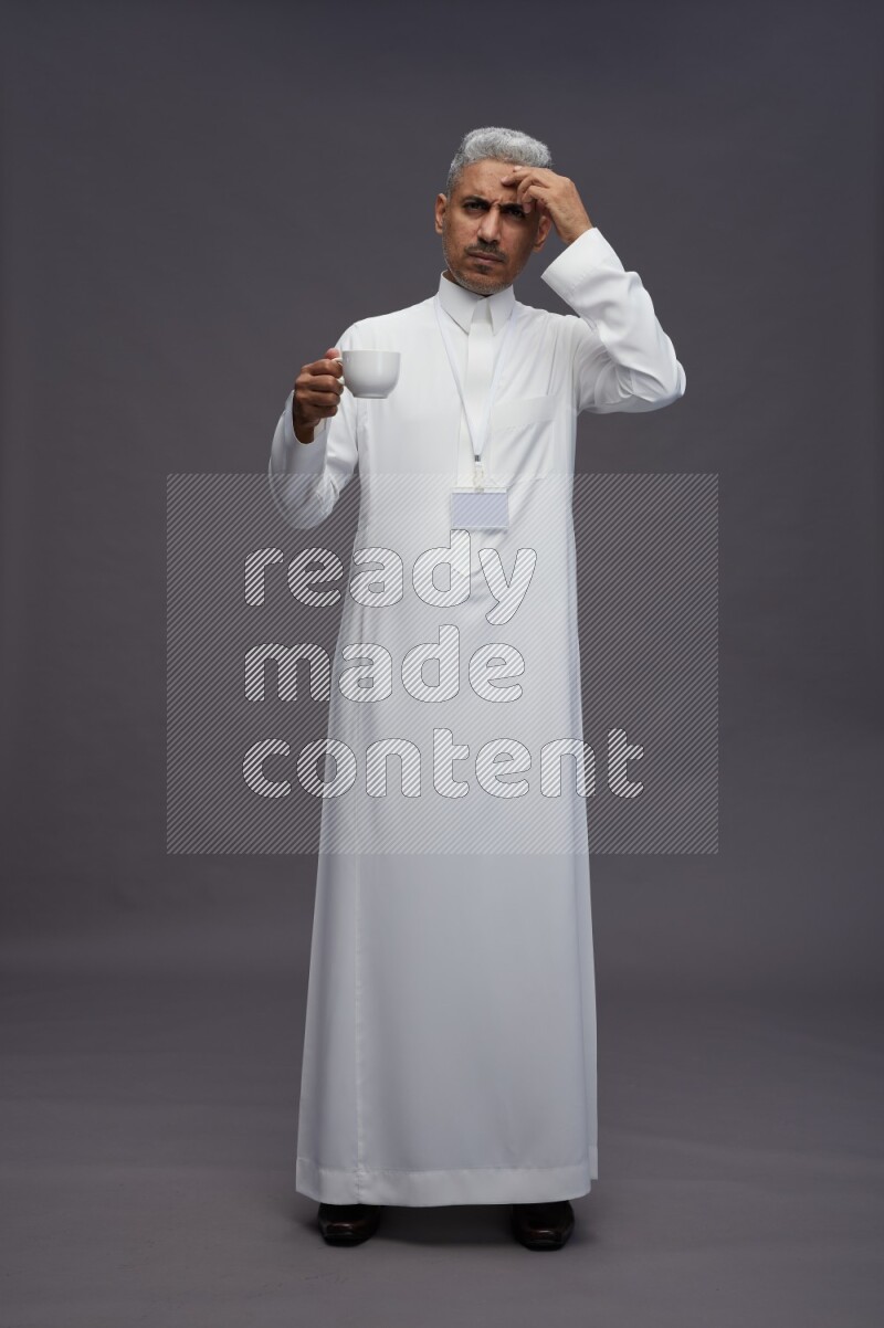 Saudi man wearing thob with neck strap employee badge standing holding mug on gray background