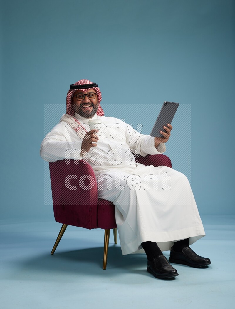 Saudi Man with shimag sitting on chair working on tablet on blue background