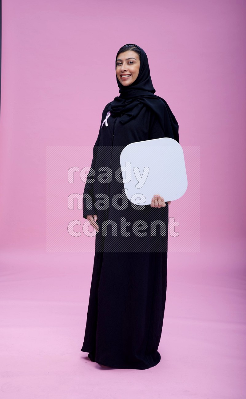 Saudi woman wearing pink ribbon on Abaya standing holding social media signs on pink background