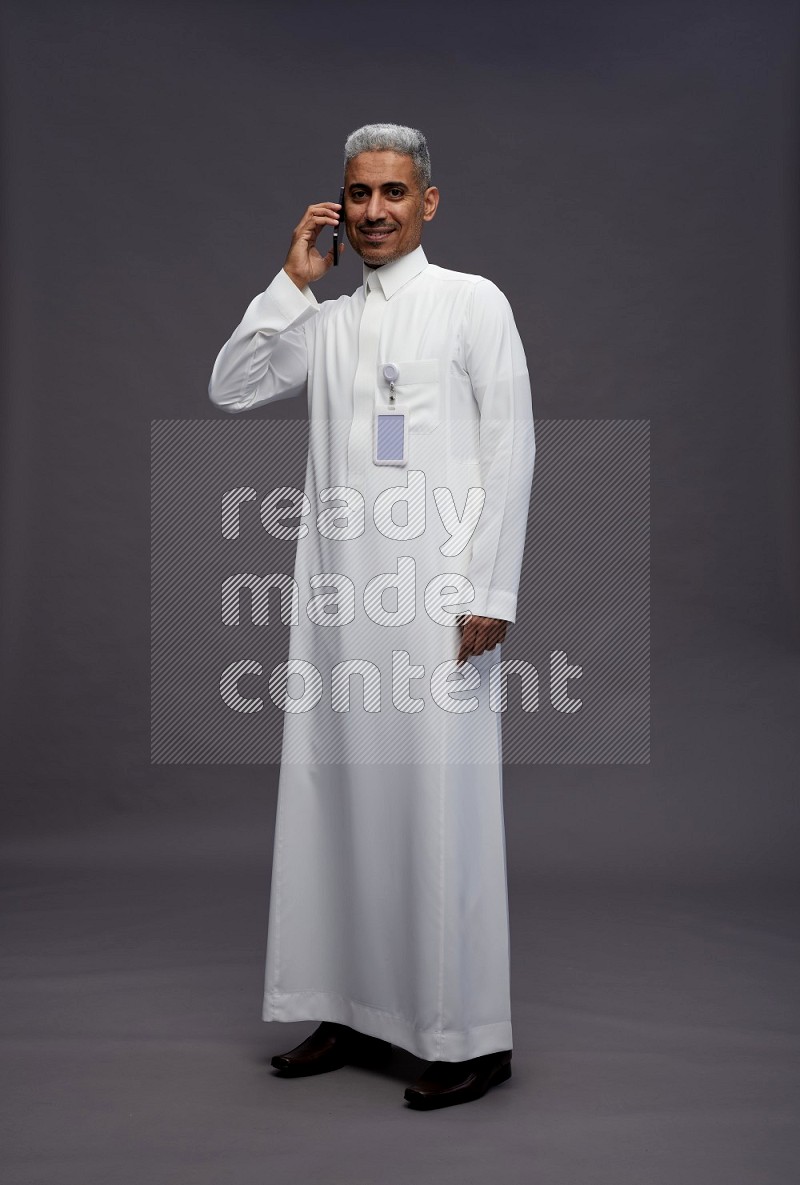 Saudi man wearing thob with pocket employee badge standing talking on phone on gray background