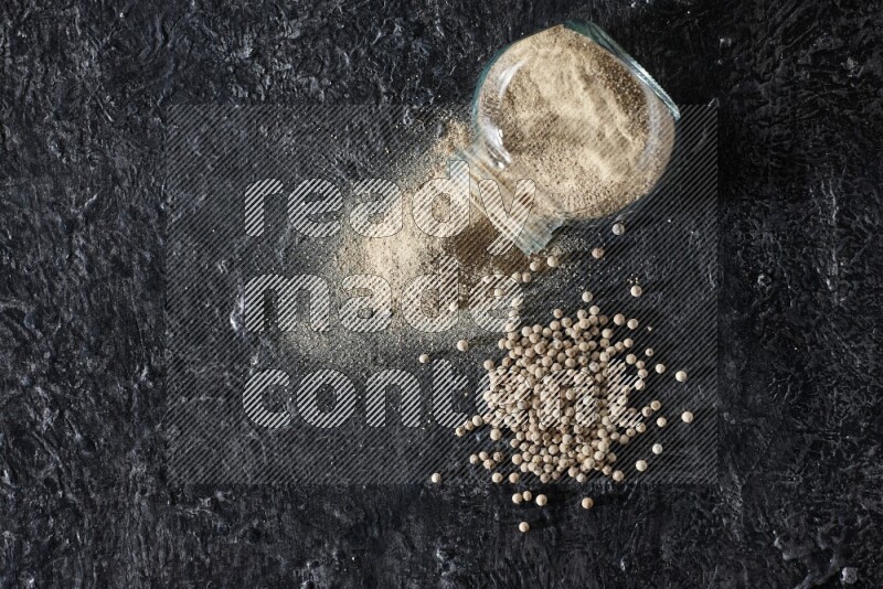 A flipped herbal glass jar full of white pepper powder with spilled powder and pepper beads on textured black flooring
