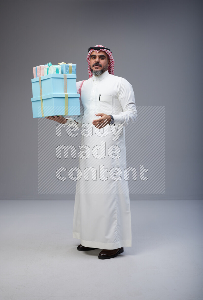 Saudi man Wearing Thob and red Shomag standing holding gift box on Gray background