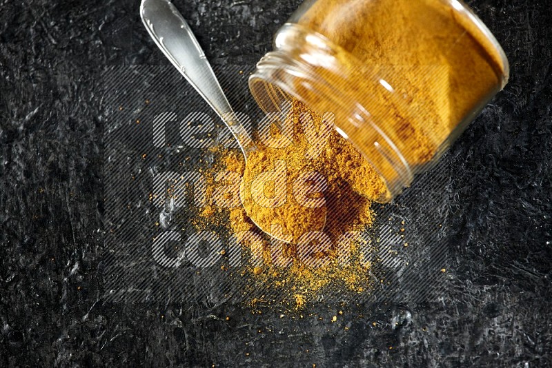 A flipped glass jar and a metal spoon full of turmeric powder and powder spilled out of it on textured black flooring