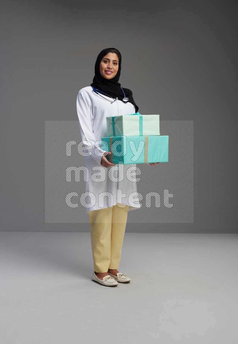 Saudi woman wearing lab coat with stethoscope standing holding gift box on Gray background