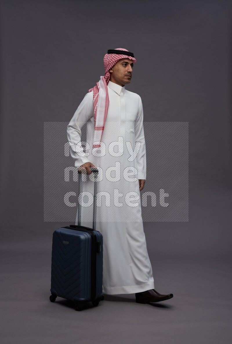 Saudi man wearing thob and shomag standing holding bag on gray background