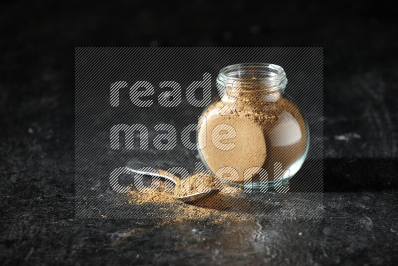A glass spice jar and metal spoon full of allspice powder on a textured black flooring