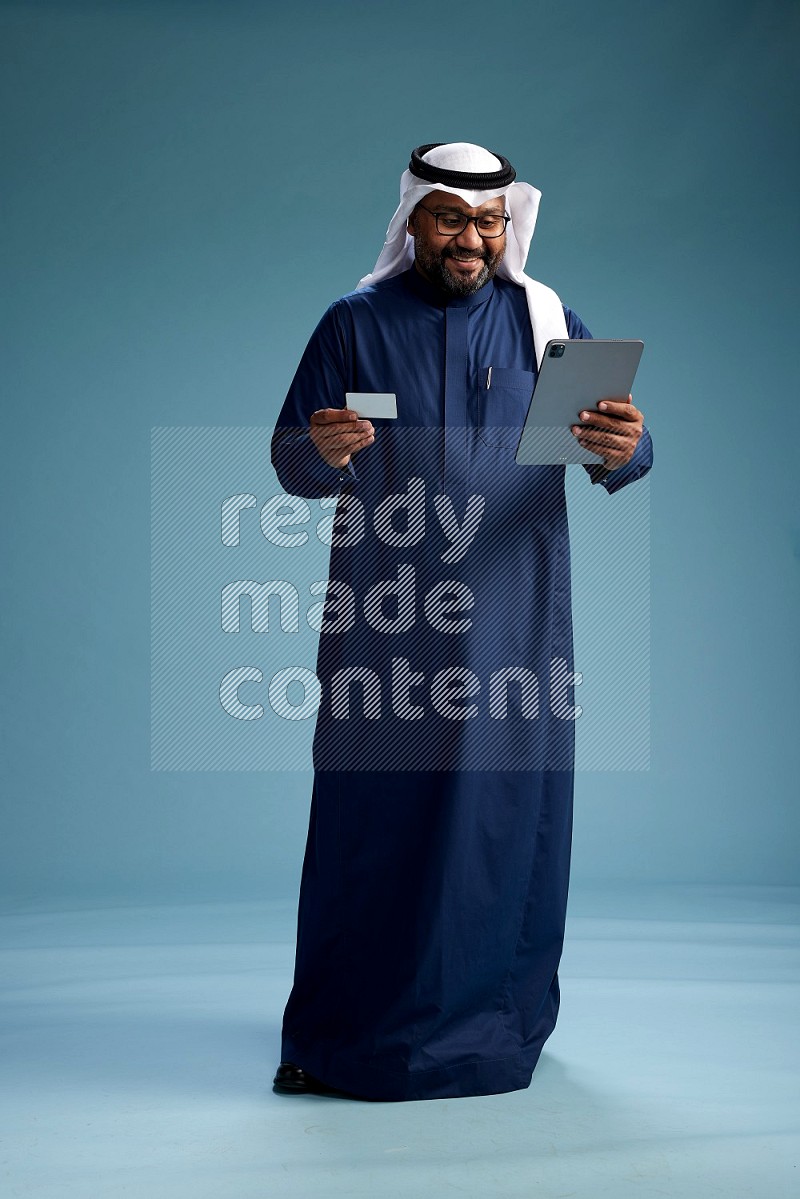 Saudi Man with shimag Standing holding ATM card while working on tablet on blue background