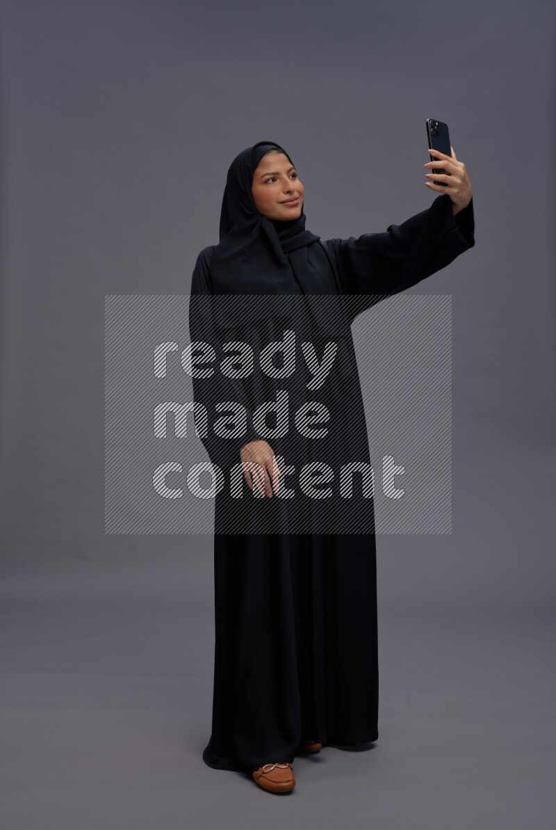Saudi woman wearing Abaya standing taking selfie on gray background