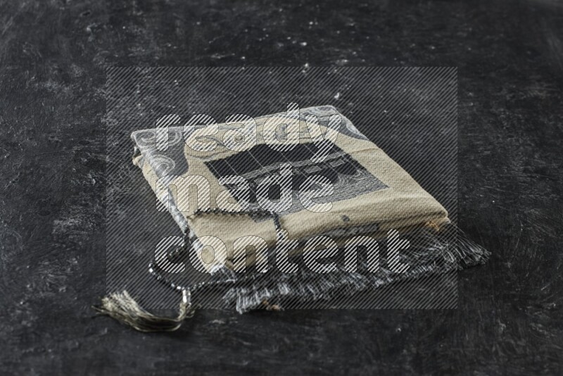 A folded prayer mat with different elements such as quran and prayer beads on black textured background