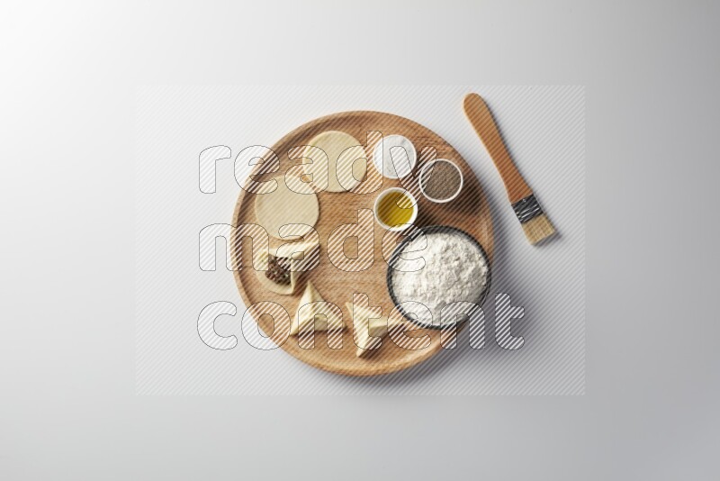 two closed sambosas and one open sambosa filled with meat while flour, salt, black pepper and oil with oil brush aside in a wooden dish on a white background