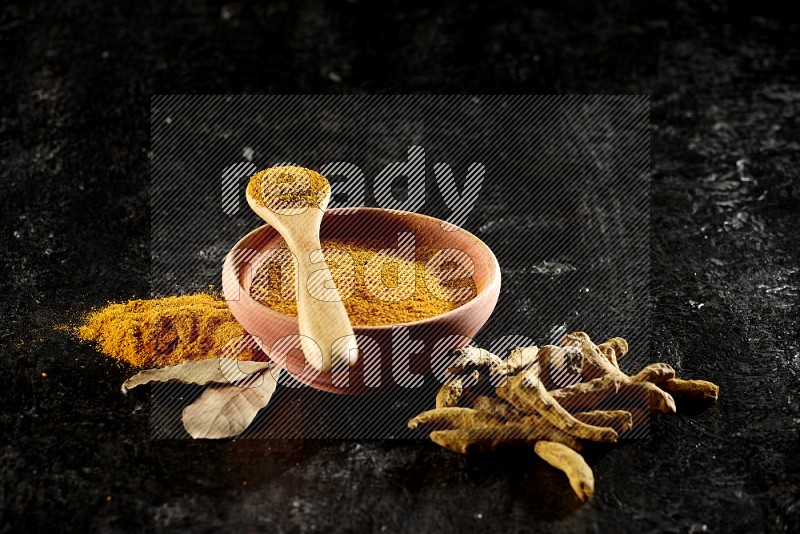 A wooden bowl and a wooden spoon full of turmeric powder with dried turmeric fingers on textured black flooring