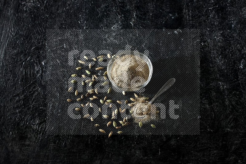 A glass bowl and a metal spoon full of cardamom powder with cardamom seeds beside them on textured black flooring
