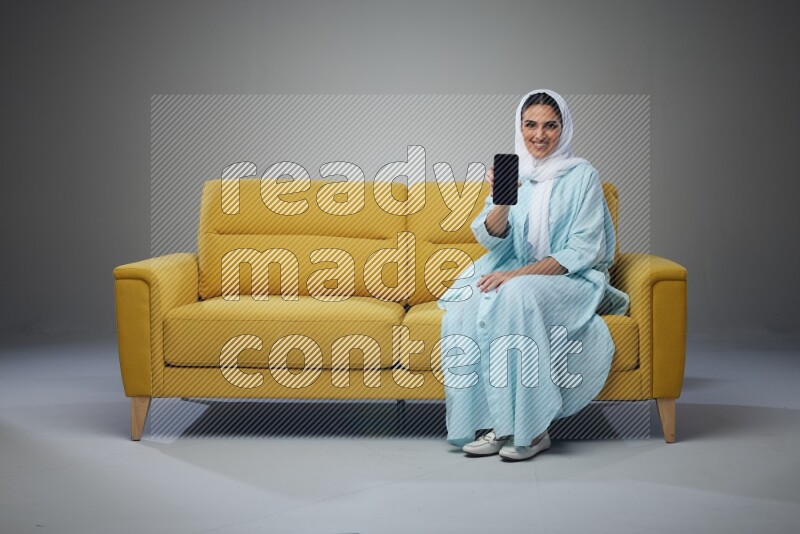 A Saudi woman wearing a light blue Abaya and white head scarf sitting on a yellow sofa and showing her phone's screen vertically and horizontally eye level on a grey background