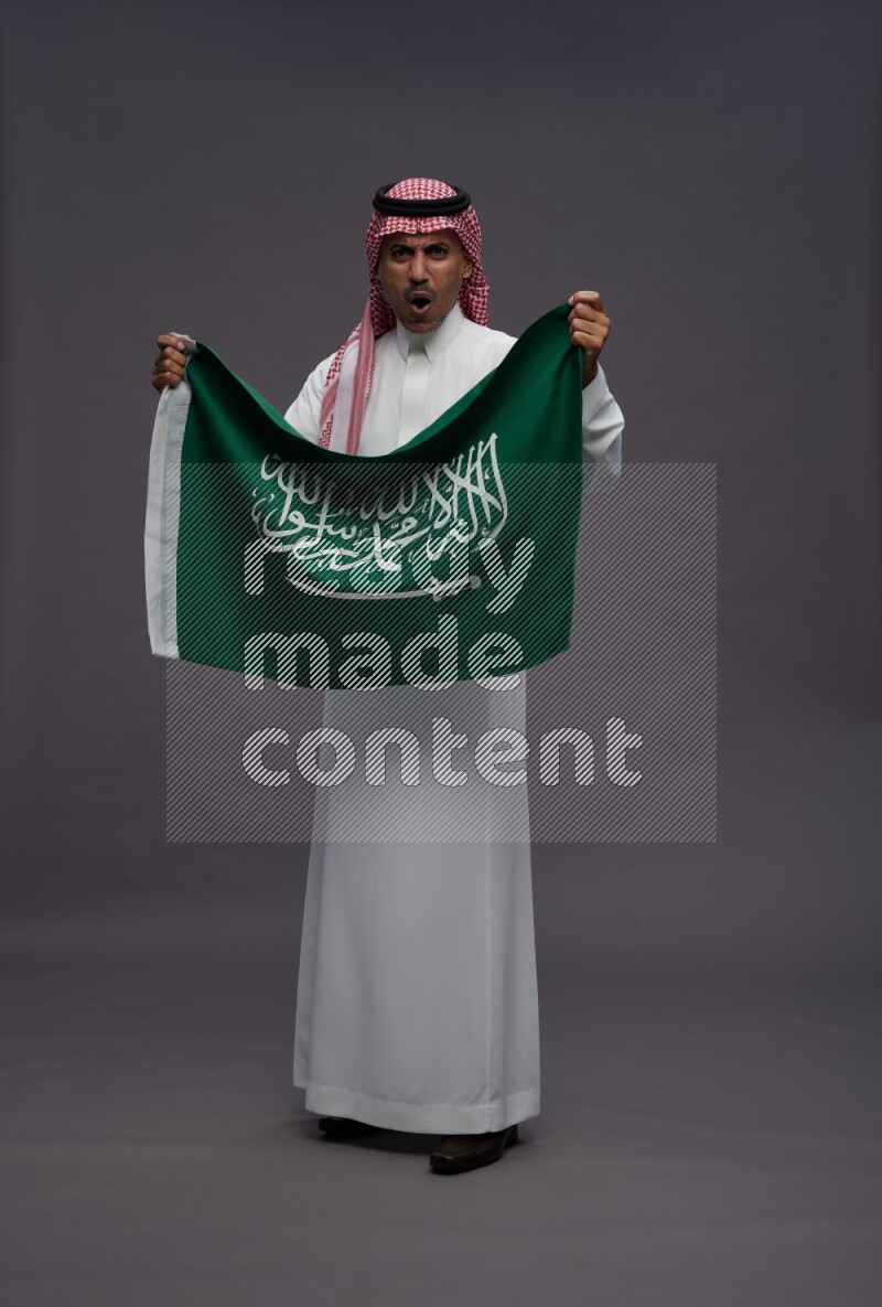 Saudi man wearing thob and shomag standing holding Saudi flag on gray background