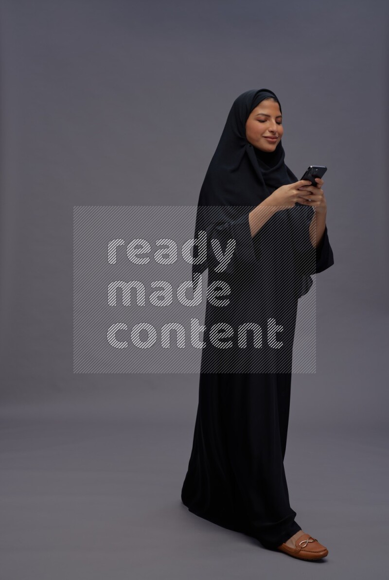 Saudi woman wearing Abaya standing texting on phone on gray background