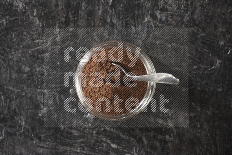 A glass jar full of cloves powder with a metal spoon on a textured black flooring