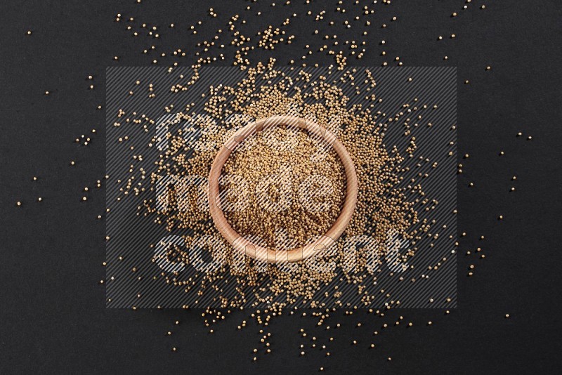 A wooden bowl full of mustard seeds and more seeds spread on a black flooring