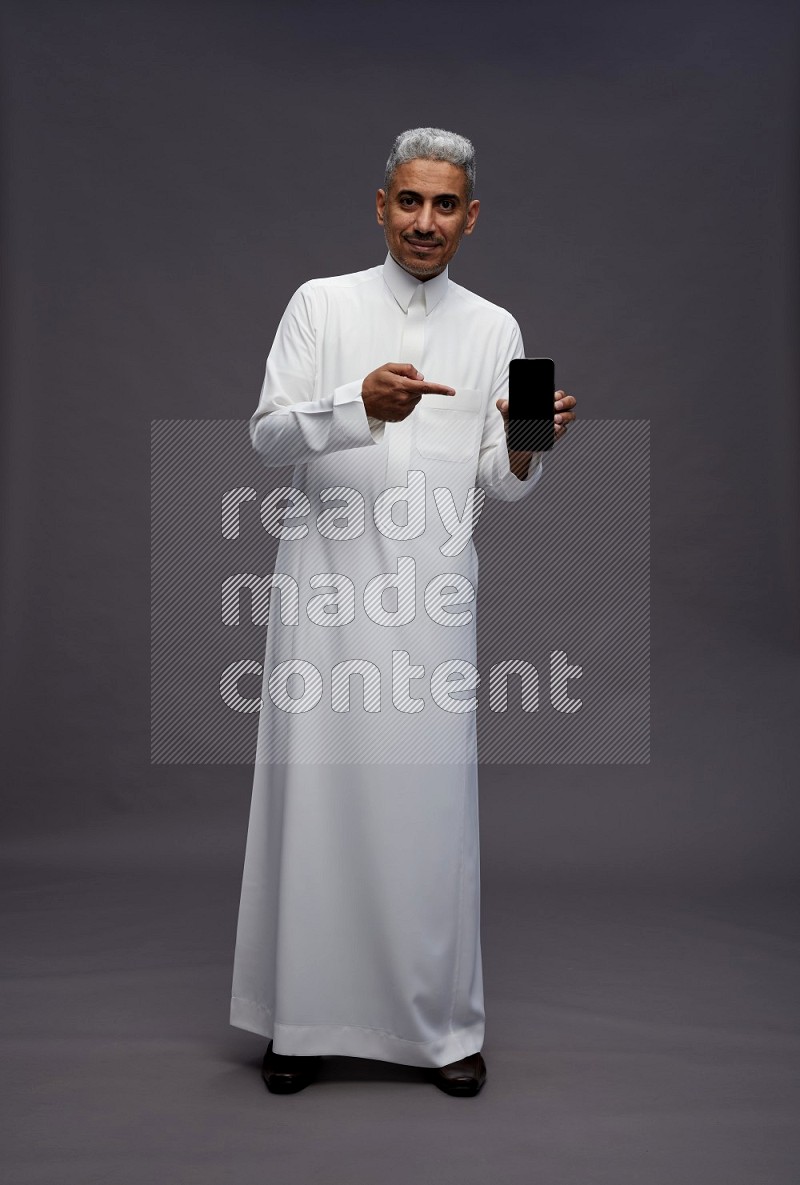 Saudi man wearing thob standing showing phone to camera on gray background