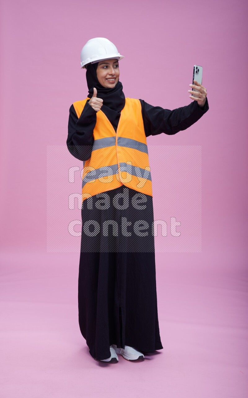 Saudi woman wearing Abaya with engineer vest and helmet standing taking selfie on pink background