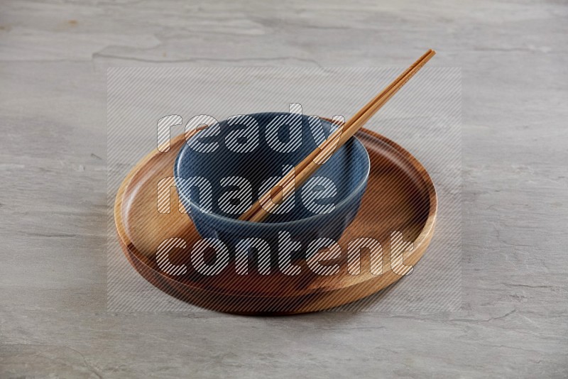 Blue pottery round bowl on top of brown wood round plate and wood chopsticks, on grey textured countertop