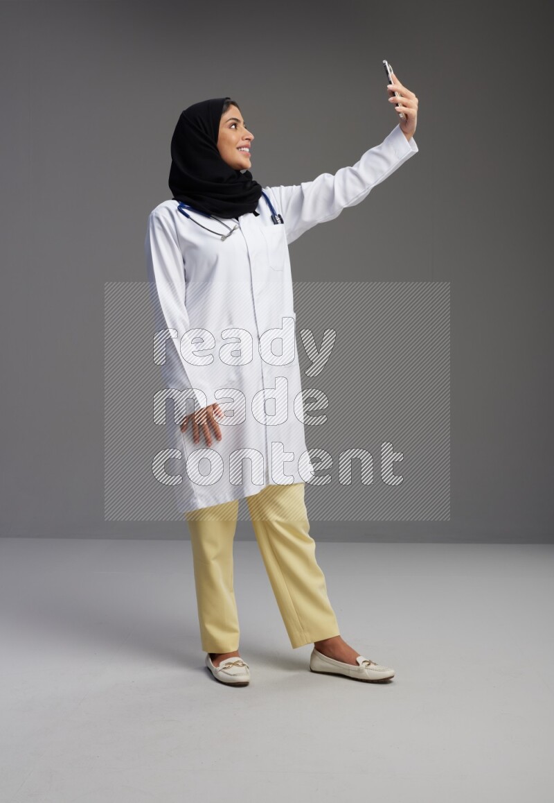 Saudi woman wearing lab coat with stethoscope standing taking selfie on Gray background