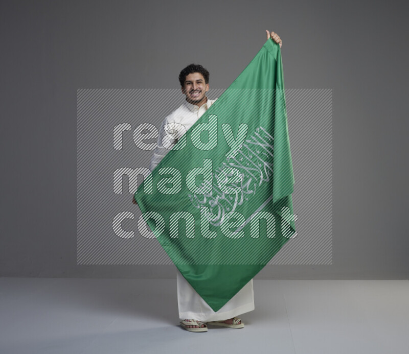 A Saudi man standing wearing thob holding big Saudi flag on gray background