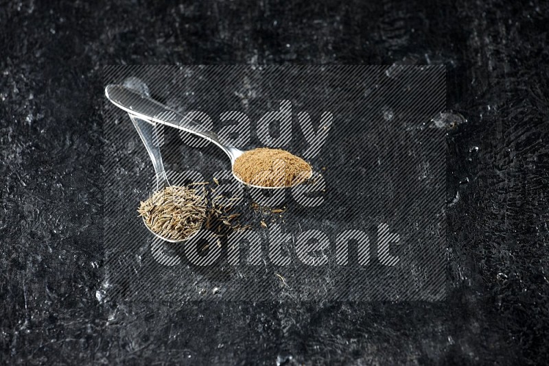 2 metal spoons full of cumin seeds and cumin powder on a textured black flooring