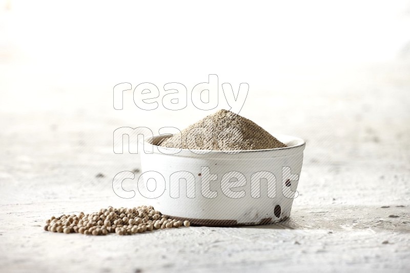 white pottery bowl full of white pepper powder with pepper beads on textured white flooring