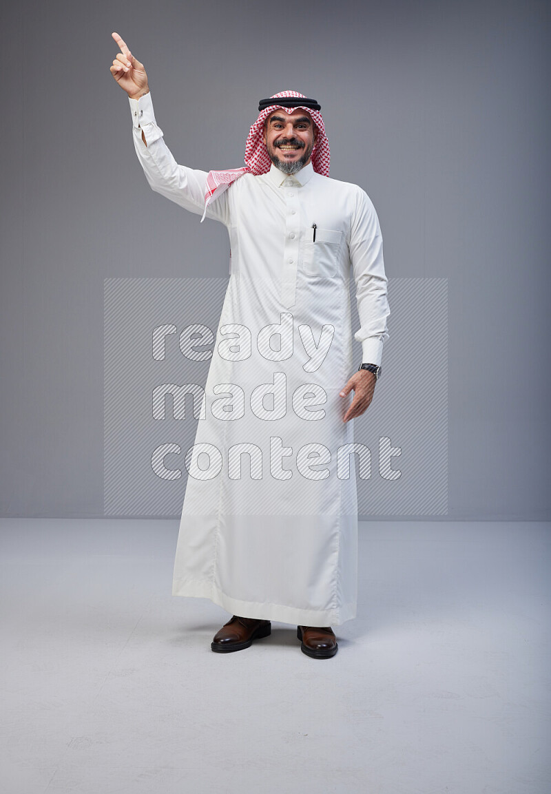 Saudi man Wearing Thob and red white Shomag standing interacting with the camera on Gray background