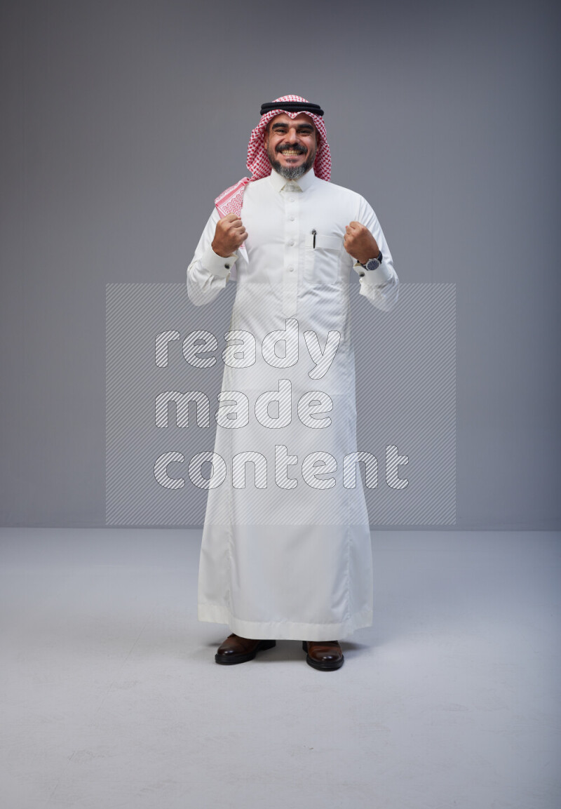 Saudi man Wearing Thob and red white Shomag standing interacting with the camera on Gray background