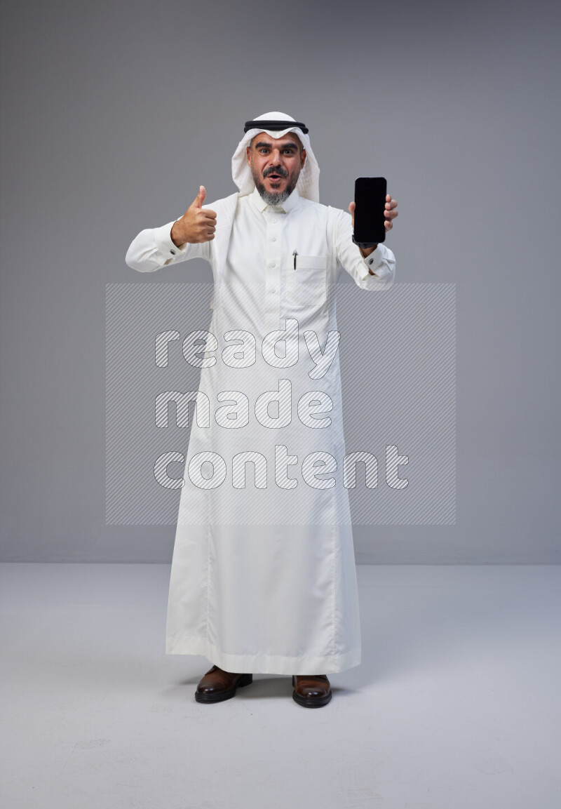 Saudi man Wearing Thob and white Shomag standing showing phone to camera on Gray background
