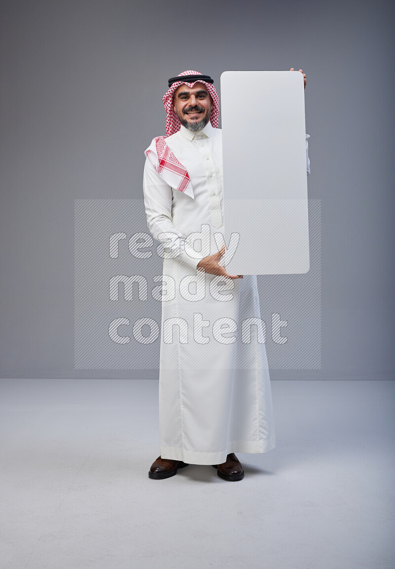 Saudi man Wearing Thob and red Shomag standing holding board on Gray background