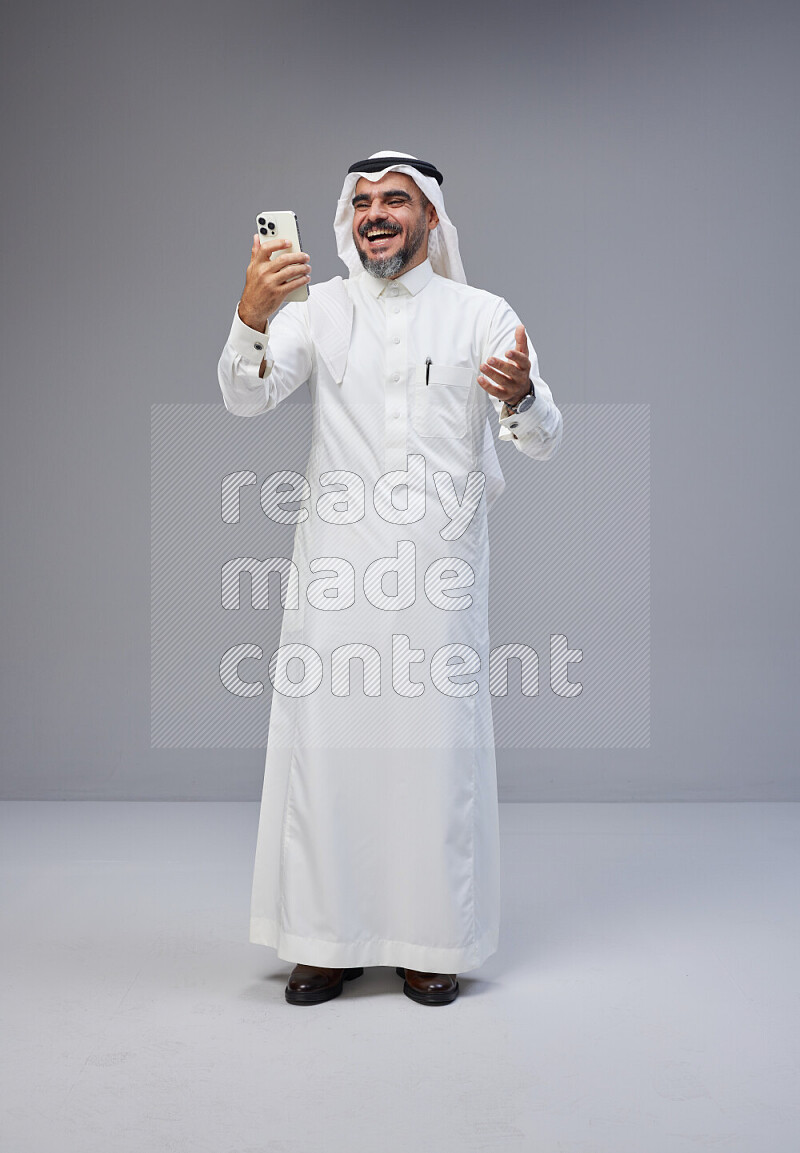 Saudi man Wearing Thob and white Shomag standing texting on phone on Gray background