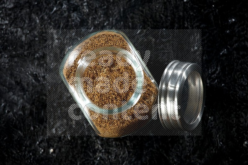 A glass spice jar full of cumin powder on a textured black flooring