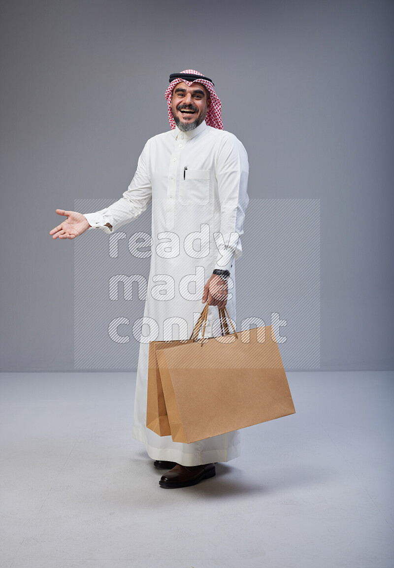 Saudi man Wearing Thob and red Shomag standing holding shopping bag on Gray background