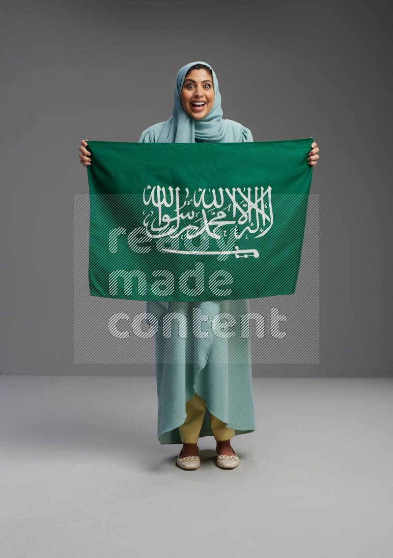 Saudi Woman wearing Abaya standing holding Saudi flag on Gray background