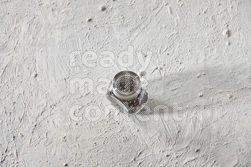 A glass spice jar full of black pepper on textured white flooring