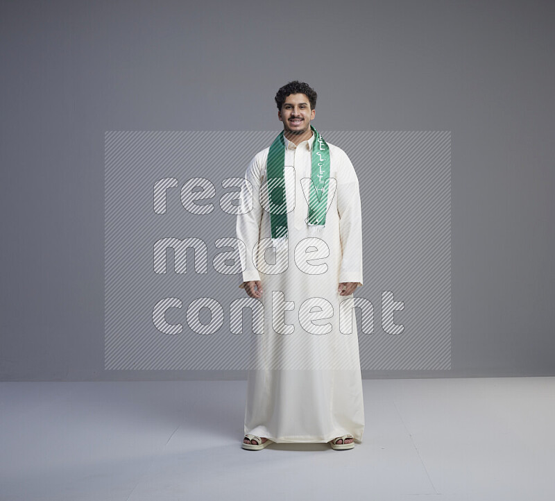 A Saudi man standing wearing thob and Saudi flag scarf on gray background