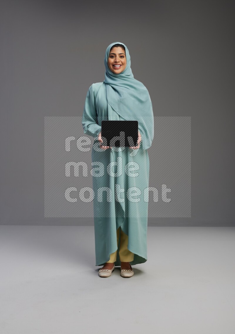 Saudi Woman wearing Abaya standing showing tablet to camera on Gray background