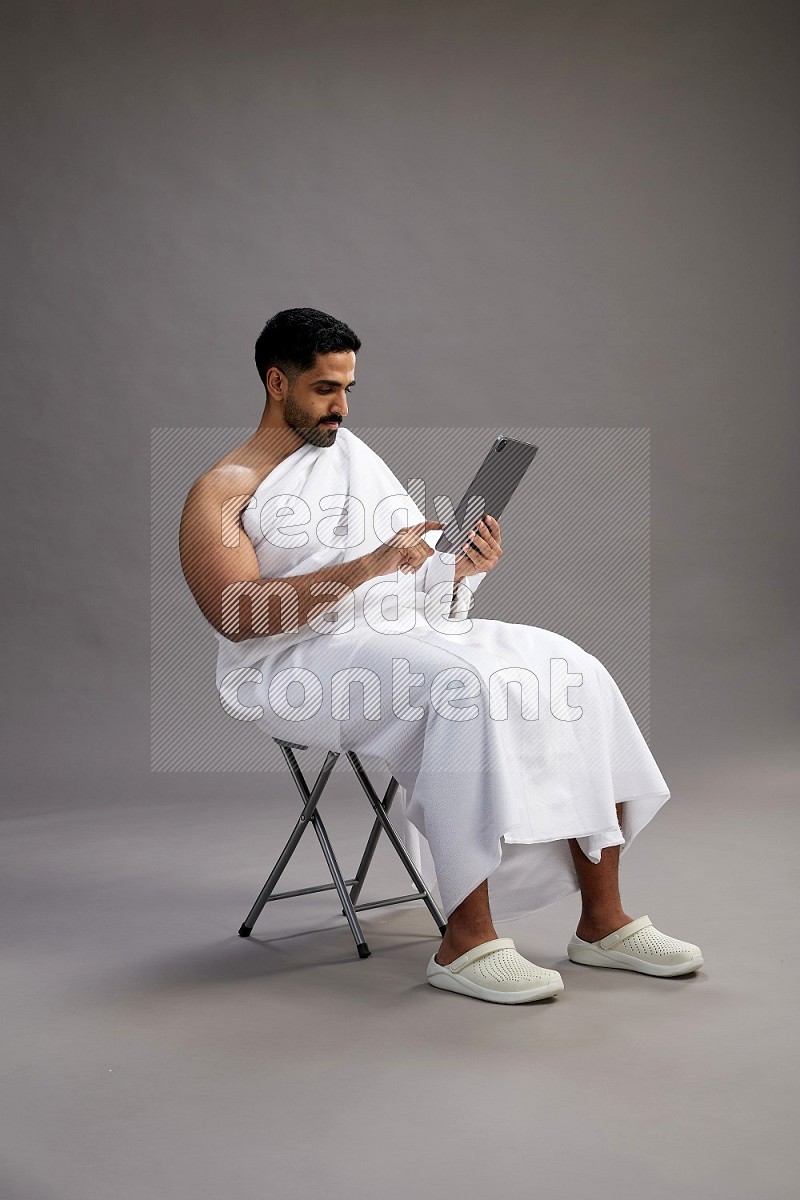 A man wearing Ehram sitting on chair holding a tablet on gray background